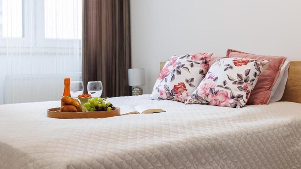 A neatly made bed with floral pillows, a tray with croissants and fruit, and a book on the bed. - Malila Apartments Łaszkiewicza 8 (Kraków)