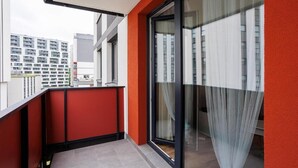 A balcony with a red wall, a glass door with curtains, and a view of other buildings. - Malila Apartments Łaszkiewicza 8 (Kraków)