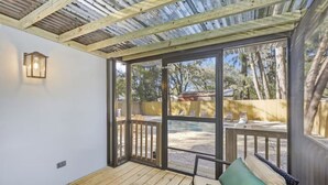 Eine überdachte Terrasse mit Holzboden, einem Rattangeflechtsofa mit Kissen, einer Laterne und großen Fenstern mit Blick auf Bäume und einen Zaun.