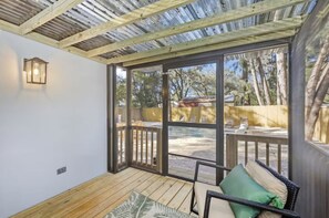 A covered patio with a wooden floor, a wicker sofa with cushions, a lantern, and large windows offering a view of trees and a fence. - NEW Saltwater Pool 3br near the beach! (Clearwater)