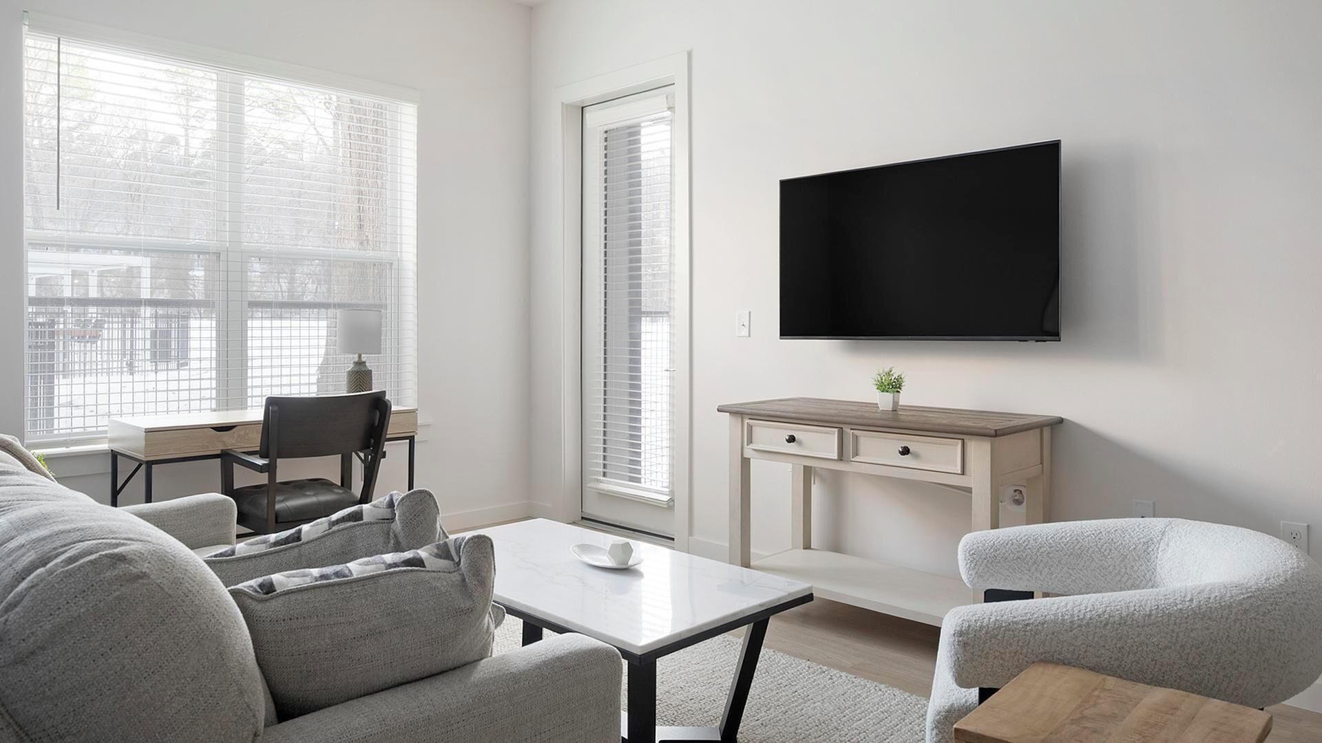 A modern living room with a sofa, armchairs, a coffee table, a TV mounted on the wall, and a desk with a chair.