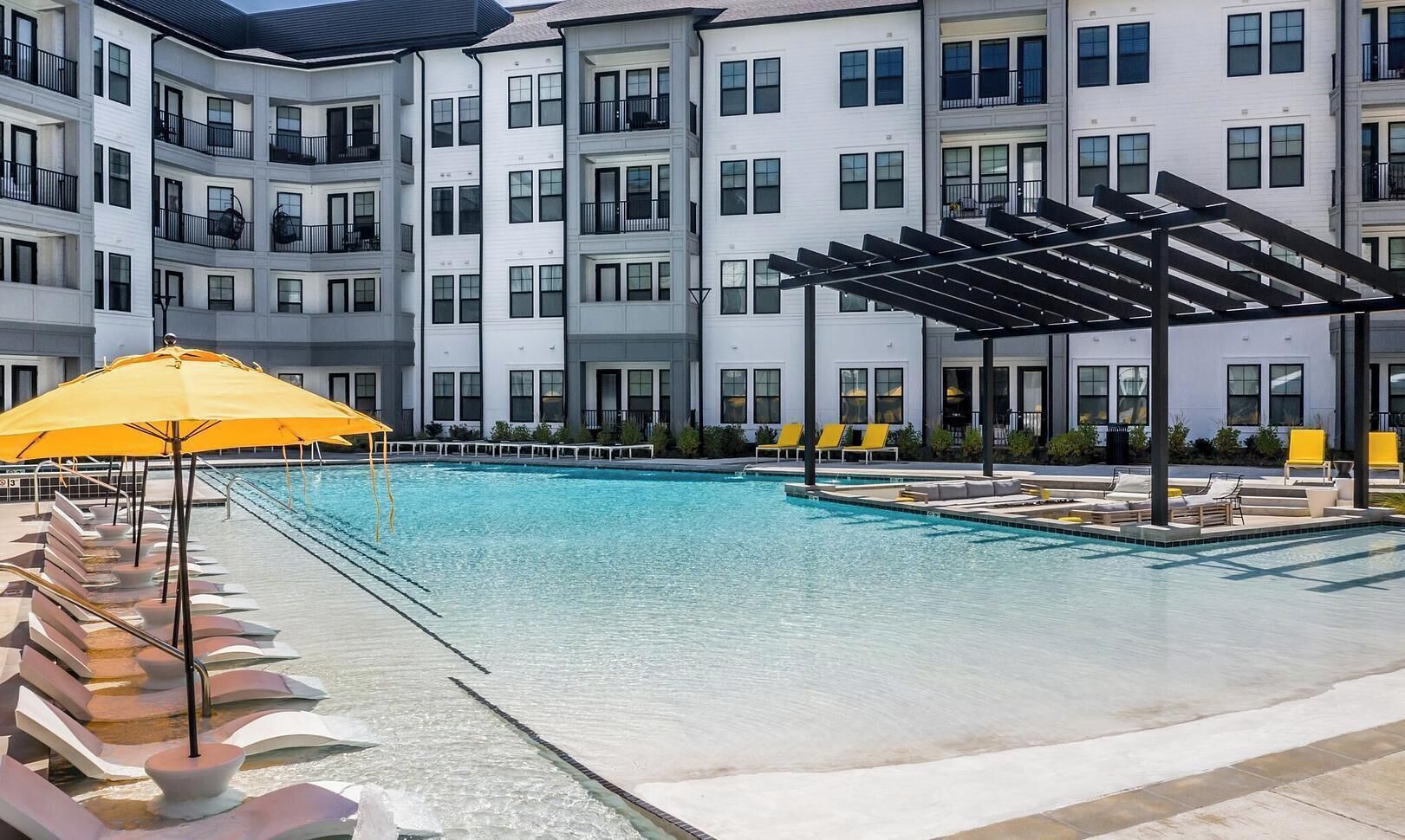 A swimming pool with lounge chairs and a yellow umbrella, surrounded by multi-story buildings.