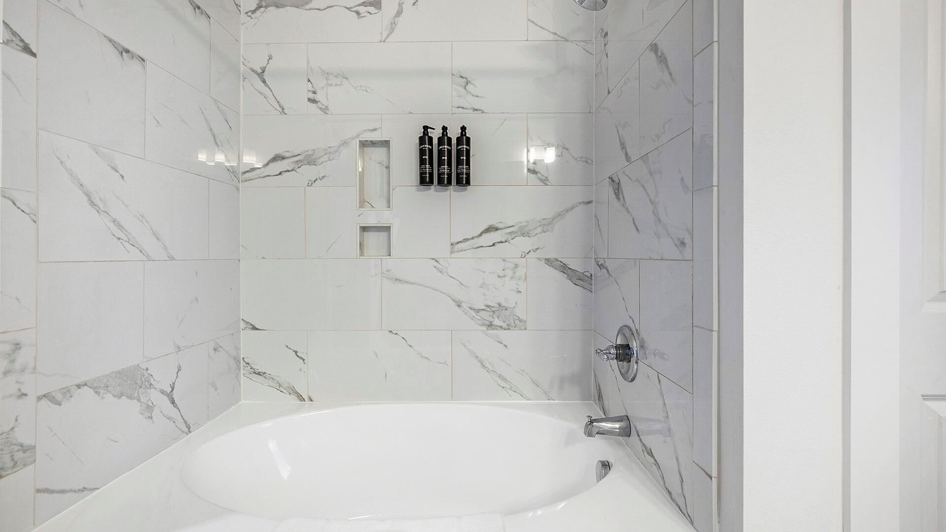 A bathroom with a marble-tiled shower wall, a white bathtub, and a shelf with toiletries.