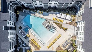 An aerial view of a modern hotel with a central swimming pool, multiple balconies, and outdoor seating areas.