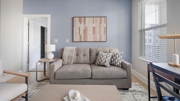 A living room with a grey sofa, a small side table with a lamp, a coffee table, and a window with blinds. - Landing | Modern 2BD, Courtyard, Gym (Little Rock)