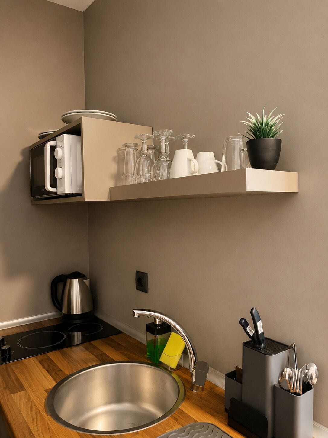 Uma cozinha moderna com uma pia de aço inoxidável, fogão embutido e uma prateleira na parede com copos e uma planta em vaso.
