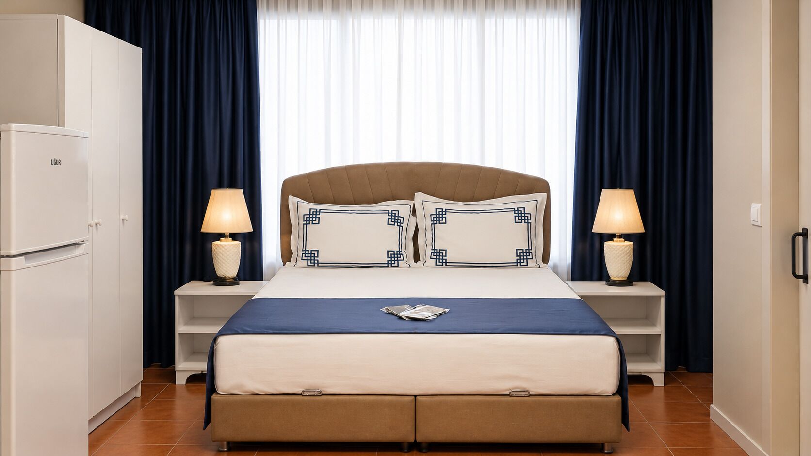 A hotel room with a bed, two bedside tables, a lamp, a wardrobe, and a window with curtains.