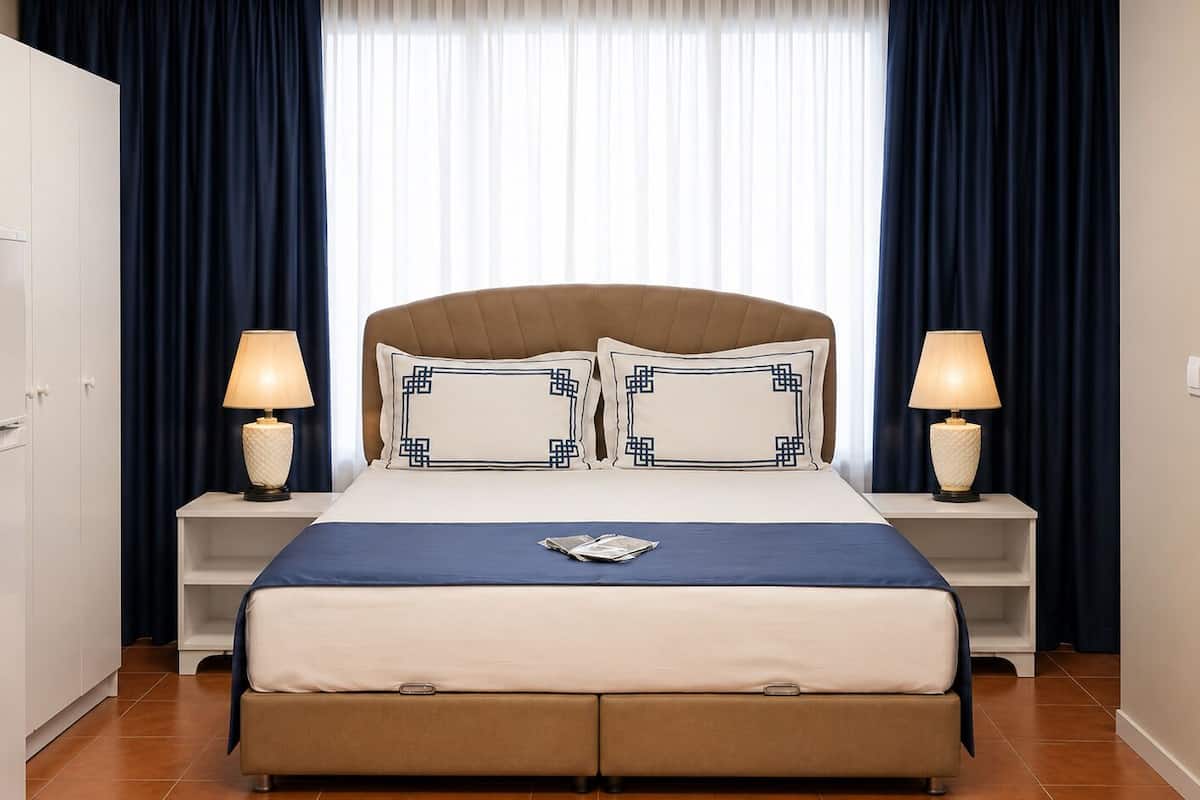 Habitación de hotel con cama, dos mesitas de noche, lámpara, armario y ventana con cortinas.