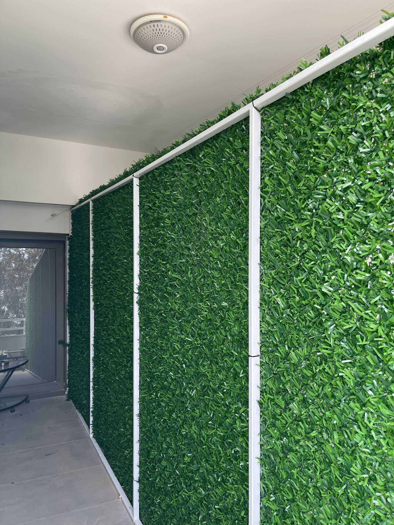 A corridor with green artificial foliage walls, a ceiling-mounted smoke detector, and a door leading to an outdoor area.