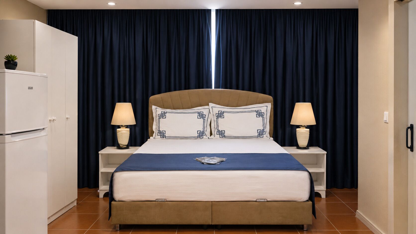 A hotel room with a bed, two bedside tables with lamps, a wardrobe, and a dark curtain backdrop.