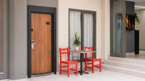 Una habitación de hotel con una puerta de madera que dice 'GO2', una puerta de cristal, una pequeña mesa con dos sillas rojas y una planta en maceta.