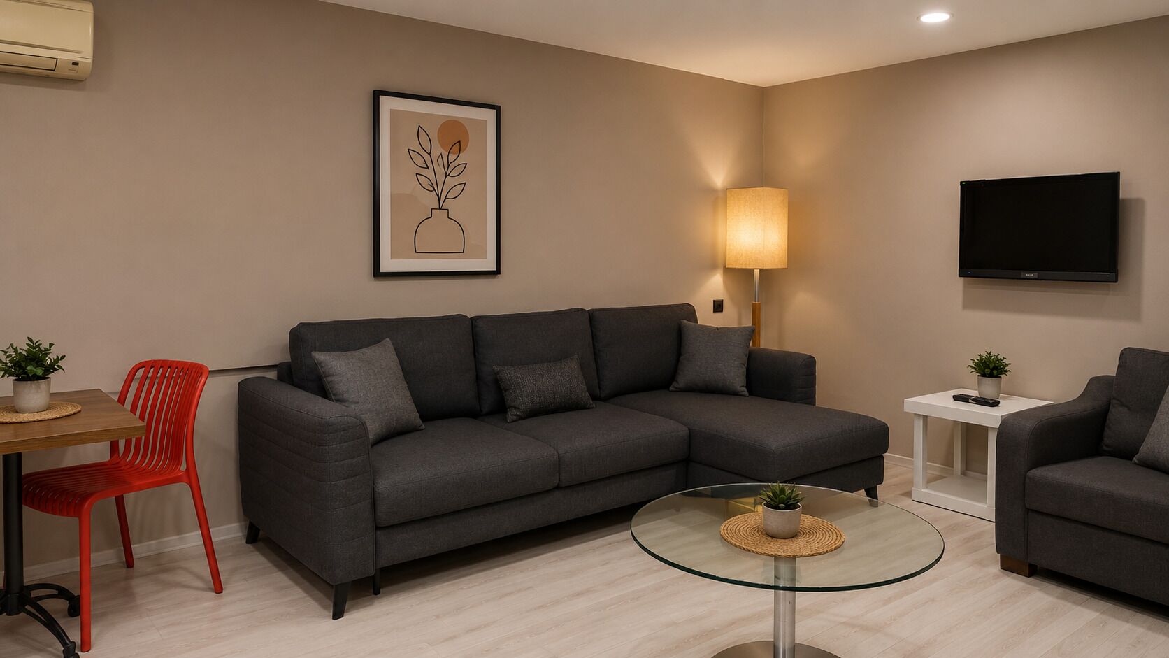 Un salon moderne avec un canapé gris, une table basse en verre, un fauteuil rouge, une petite table avec une plante, un téléviseur fixé au mur et un tableau encadré accroché au mur.