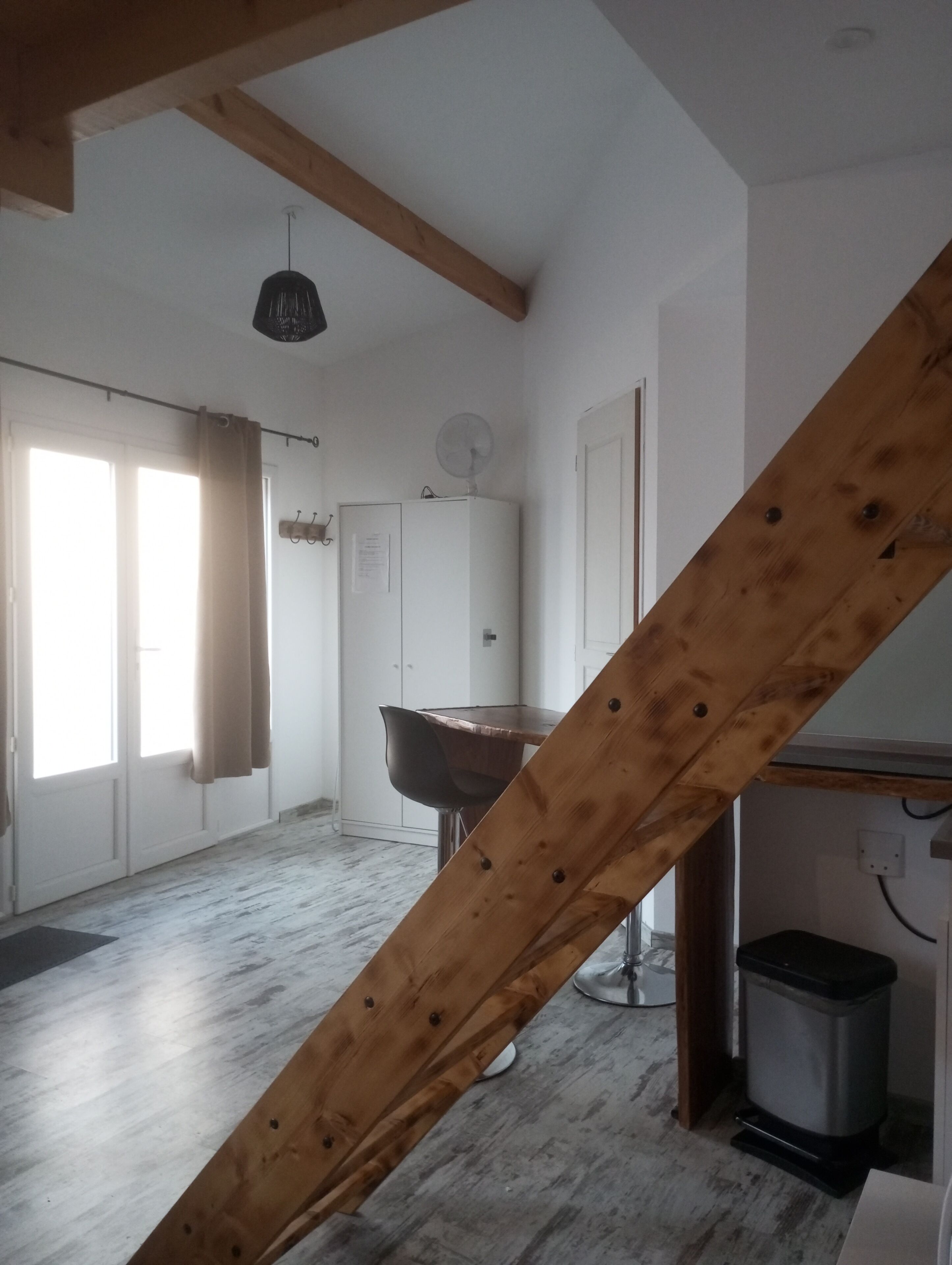 A room with a wooden ceiling beam, a dining table with chairs, a refrigerator, a window with curtains, and a trash bin.