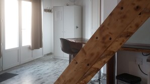 A room with a wooden ceiling beam, a dining table with chairs, a refrigerator, a window with curtains, and a trash bin.