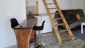 A loft-style room with a wooden ladder, a dining table with chairs, a kitchenette, and a bed with a patterned rug.
