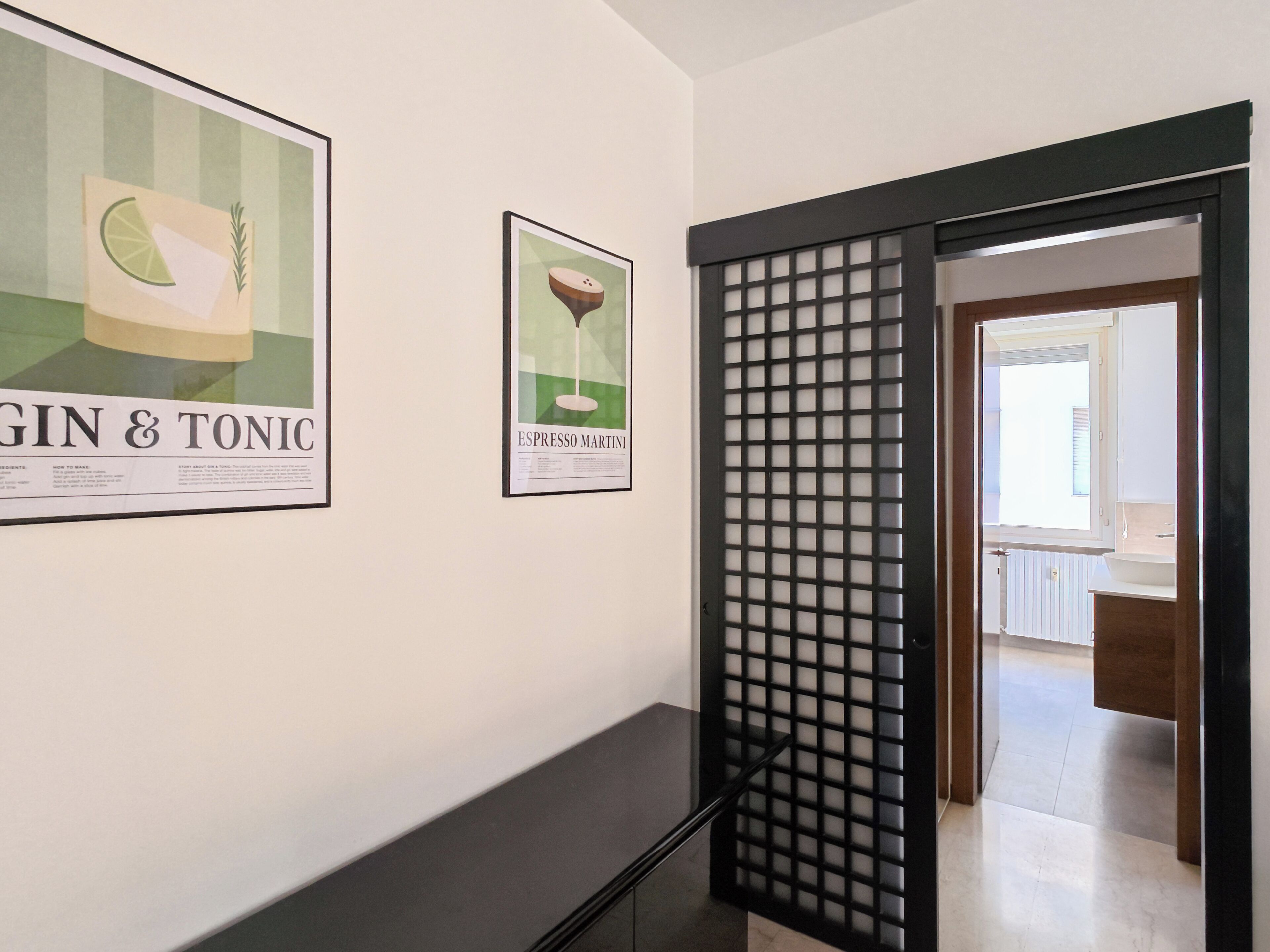 Un pasillo con carteles enmarcados en la pared, un separador negro y una puerta abierta que da a un baño.