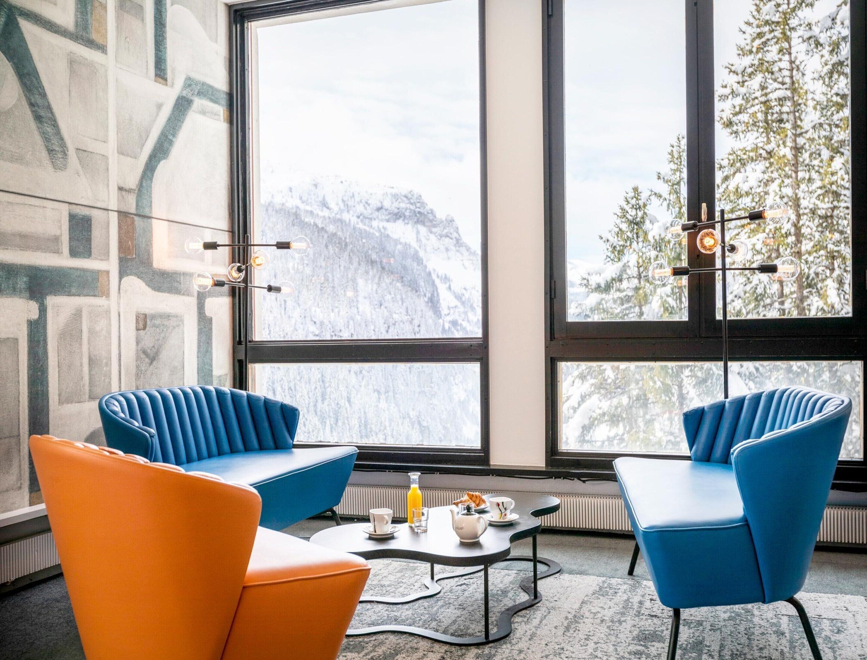 A modern room with large windows, two blue armchairs, an orange chair, a coffee table, and a radiator.