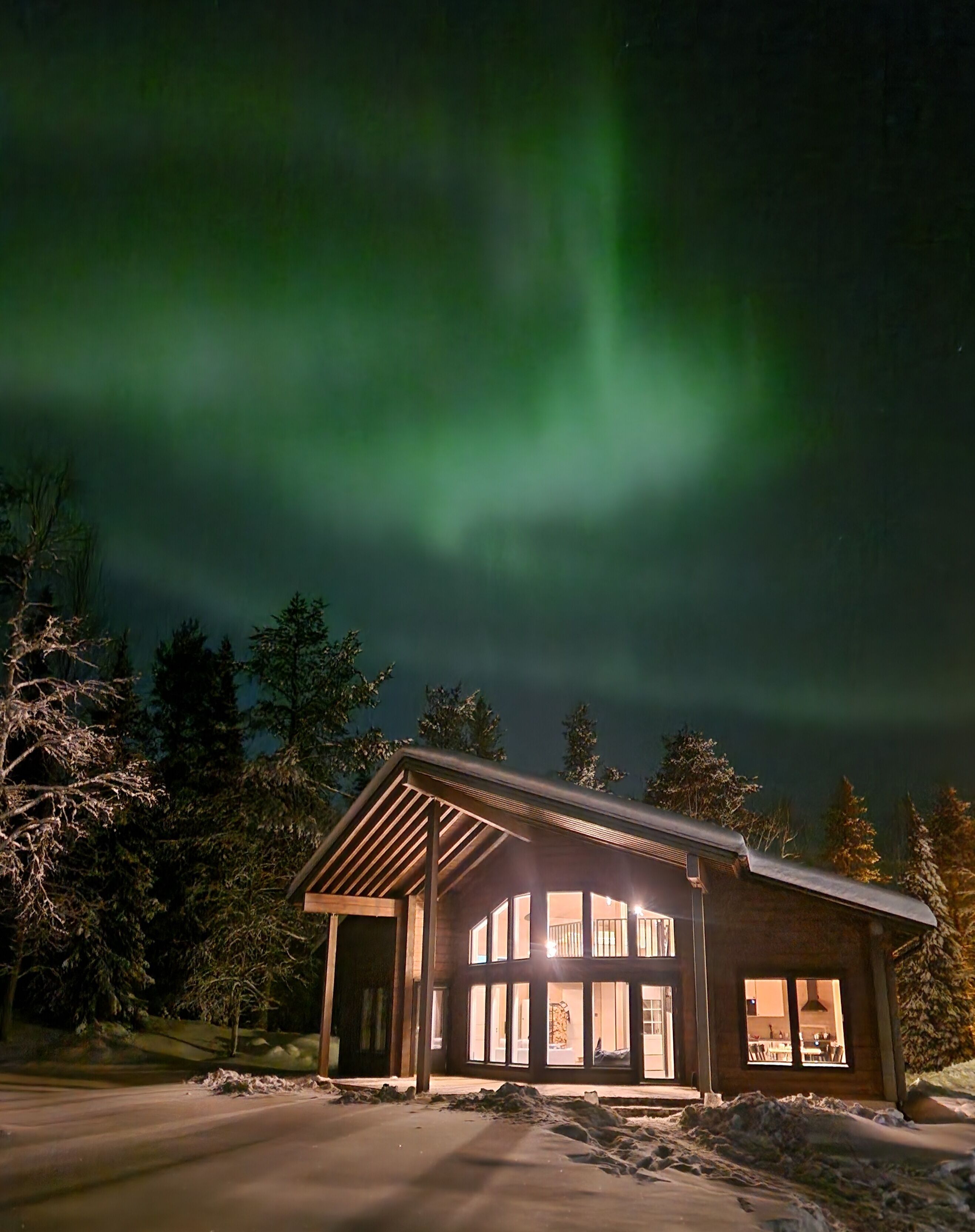 Eine Lodge mit großem Fensterfront, umgeben von Schnee und Bäumen, unter einem Nachthimmel mit sichtbaren Polarlichtern.