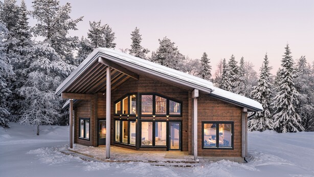 Puinen mökki, jossa on suuret ikkunat, katettu kuisti ja lumipeitteinen maisema.