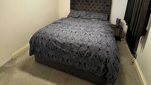 A bedroom with a tufted headboard, a patterned bedspread, a bedside table with a lamp, and a window with curtains. - Charming Bungalow Rental in Rainham Comfort and Convenience (Rainham)