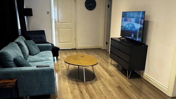 A living room with a sofa, coffee table, TV stand, and a clock on the wall. - Charming Bungalow Rental in Rainham Comfort and Convenience (Rainham)