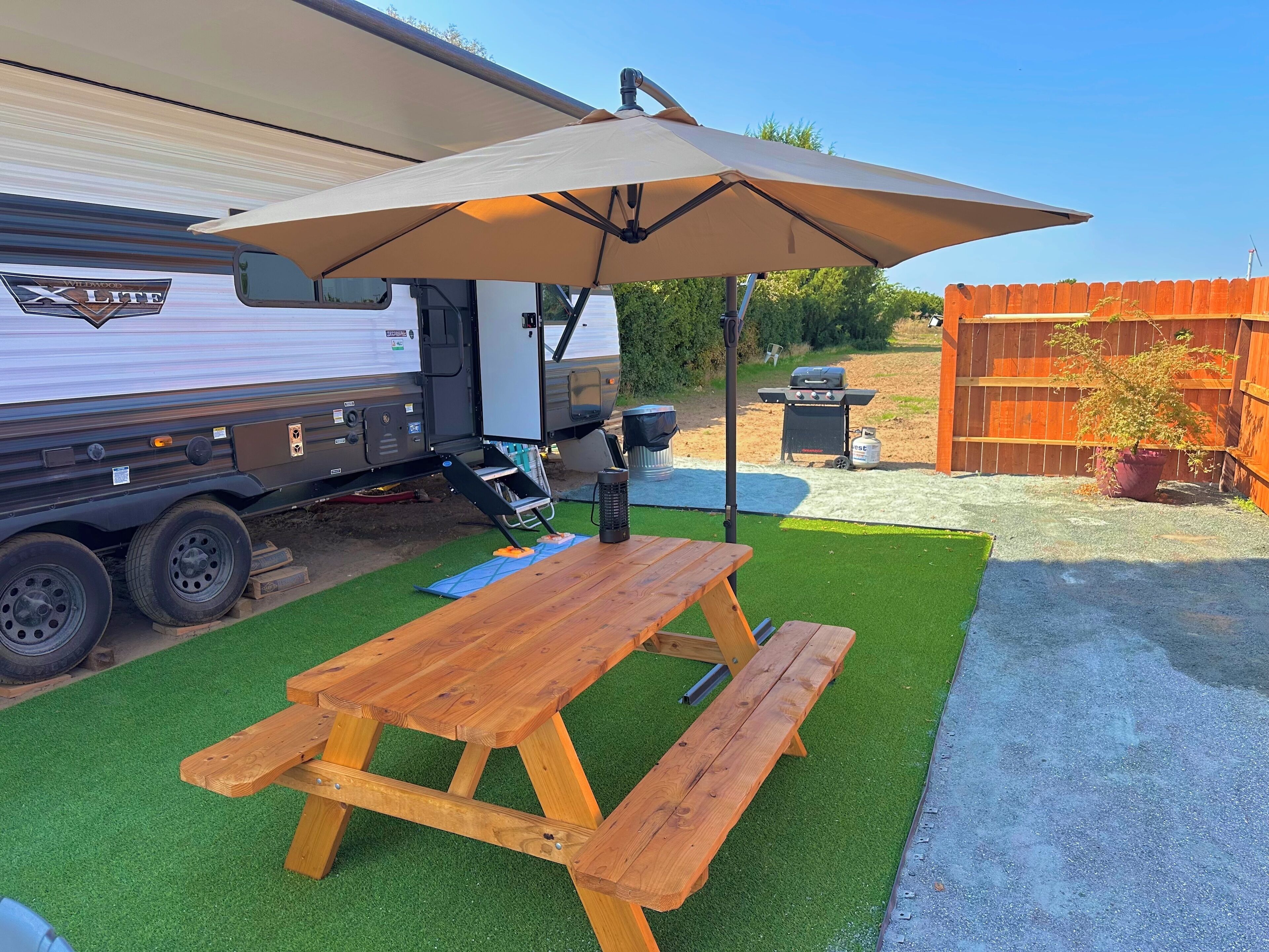 En campingplats med ett picknickbord, ett parasoll, en grill och en husvagn.