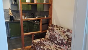 A living area with a sofa, wooden shelves, a kitchen in the background, and a herringbone-patterned floor.