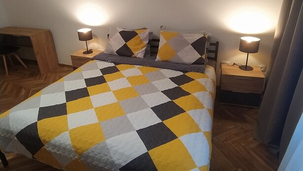 A bed with a checkered bedspread, two bedside lamps, a wooden nightstand, a desk, and a painting on the wall.