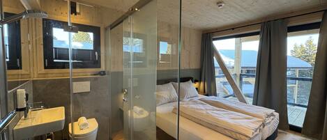A modern bedroom with a glass shower enclosure, a large bed, a built-in sink, and a view of the outdoors.