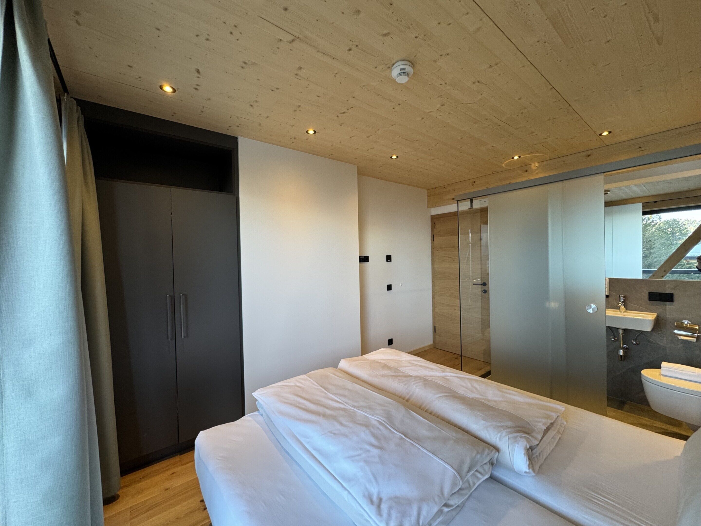 A modern bedroom with a wooden ceiling, a large bed with white bedding, a walk-in closet, and a bathroom with a mirror and sink.