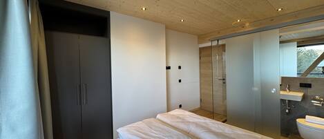 A modern bedroom with a wooden ceiling, a large bed with white bedding, a walk-in closet, and a bathroom with a mirror and sink.