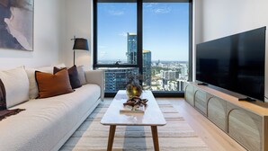 A modern living room with a large window, a sofa, a coffee table, and a flat-screen TV. - City Lights & Designer Comfort w Heated Pool, Gym, Sauna, Steam, Bar and more (Southbank)