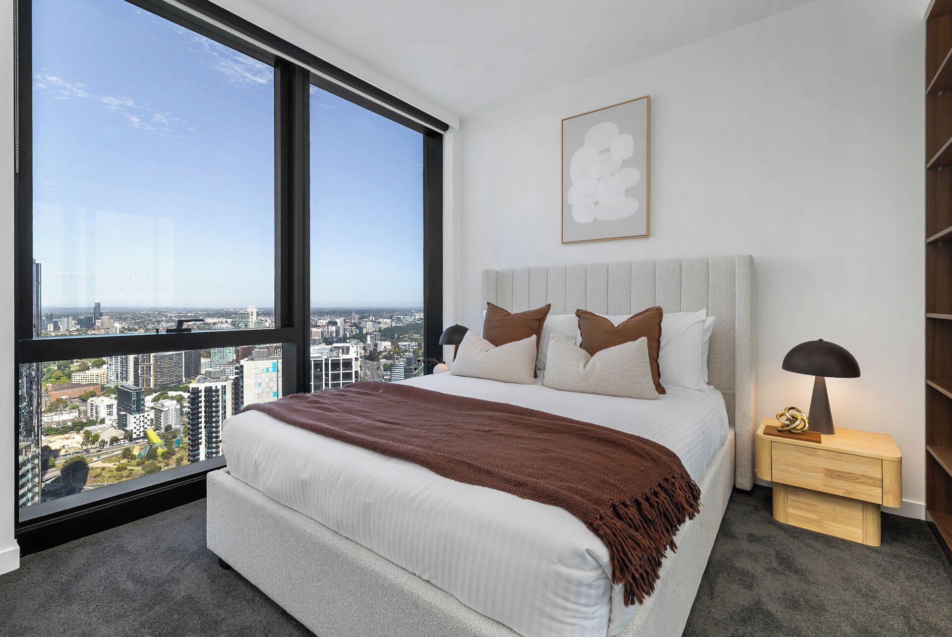 A bedroom with a large window, a bed with pillows, a bedside table, and a wall-mounted artwork.