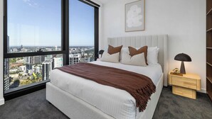 A bedroom with a large window, a bed with pillows, a bedside table, and a wall-mounted artwork. - City Lights & Designer Comfort w Heated Pool, Gym, Sauna, Steam, Bar and more (Southbank)