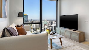 A modern living room with a large window, a flat-screen TV, a sofa, a coffee table, and a cabinet. - City Lights & Designer Comfort w Heated Pool, Gym, Sauna, Steam, Bar and more (Southbank)