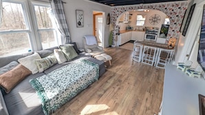A cozy living room with a sofa, a dining area with a table and chairs, a kitchen with a stove, and a large window with curtains. - Dogs Stay Free! Lakefront Home near Notre Dame (South Bend)