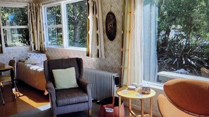 A living room with a sofa, armchair, and a small table. There are large windows with curtains, a radiator, and a view of trees outside. - A short walk to the beautiful Lake Taupo relax and take in the fabulous views! (Kuratau)