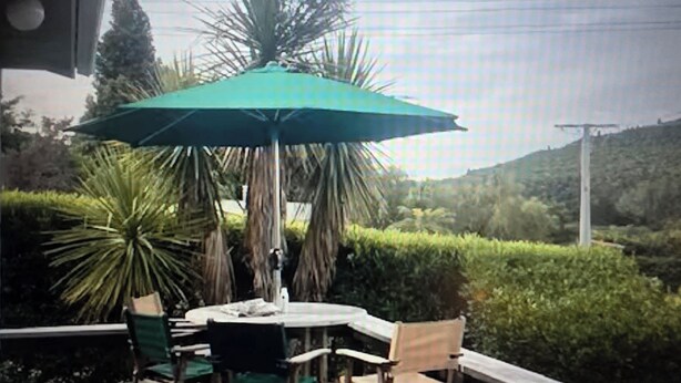 A wooden deck with a table and chairs under a green umbrella, surrounded by greenery and hills. - A short walk to the beautiful Lake Taupo relax and take in the fabulous views! (Kuratau)