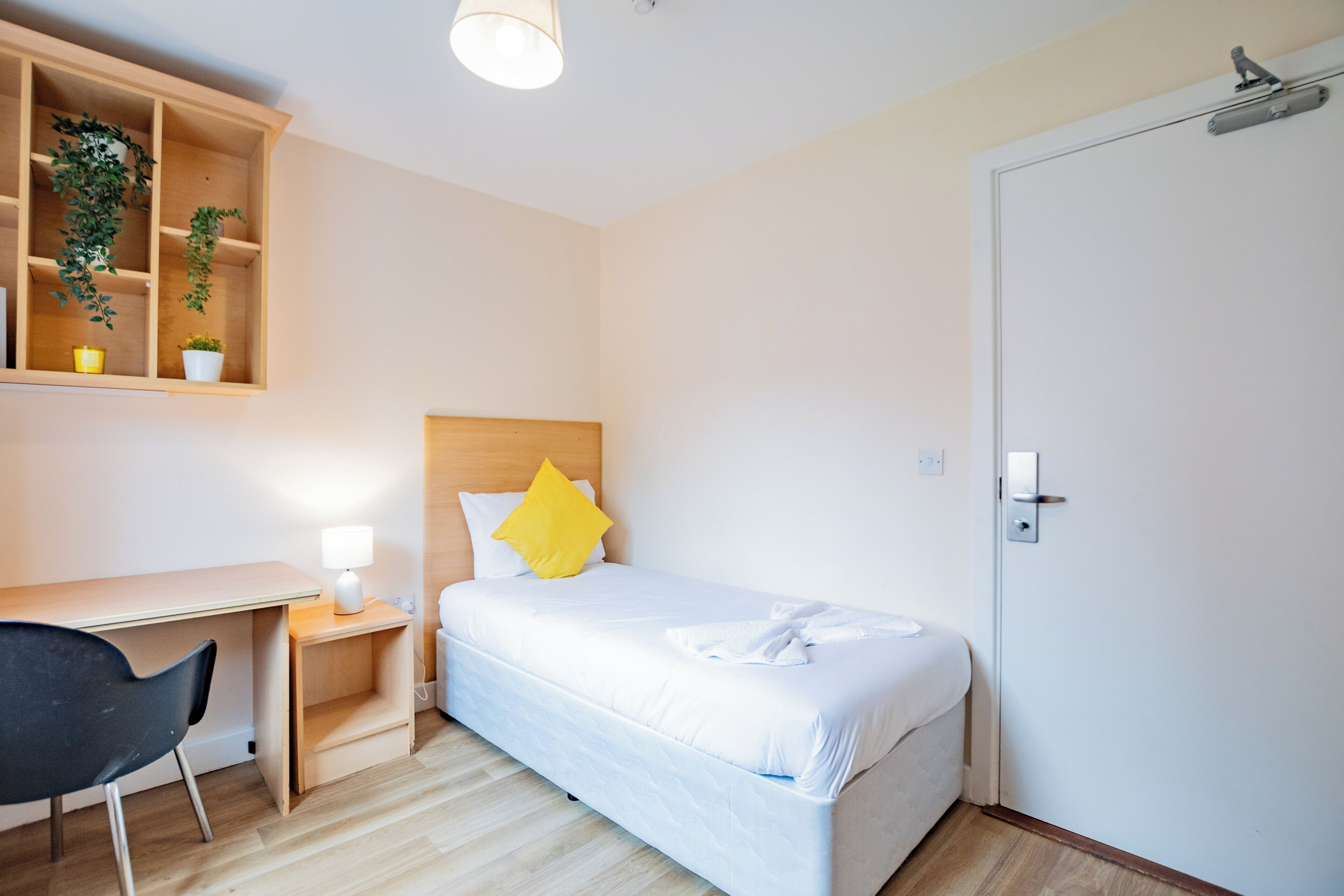 Habitación de hotel con cama, escritorio, silla, mesita de noche, lámpara y puerta.