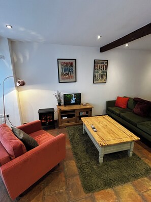 Un salón con un sofá rojo, un sofá verde, una mesa de centro de madera, un televisor sobre un mueble de madera y dos carteles enmarcados en la pared.