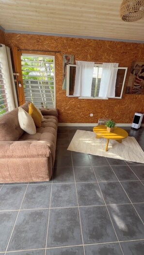 A living room with a sofa, a small table, a potted plant, and a window with curtains. - LAKAZ'DELAIR - Quiet bungalow with Jacuzzi, close to town and beaches. (Sainte-Anne)