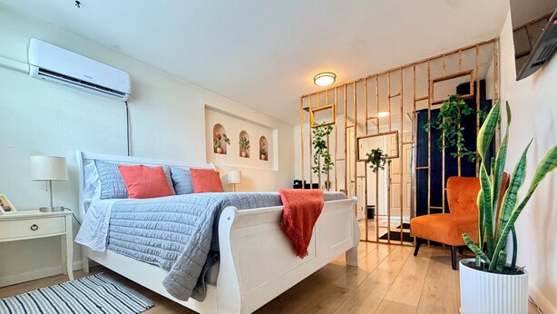 A bedroom with a bed, bedside table, lamp, air conditioning unit, potted plant, and a chair. - Sunny Baldwin Park Studio • Walk to Cafés • Near Downtown (Orlando)