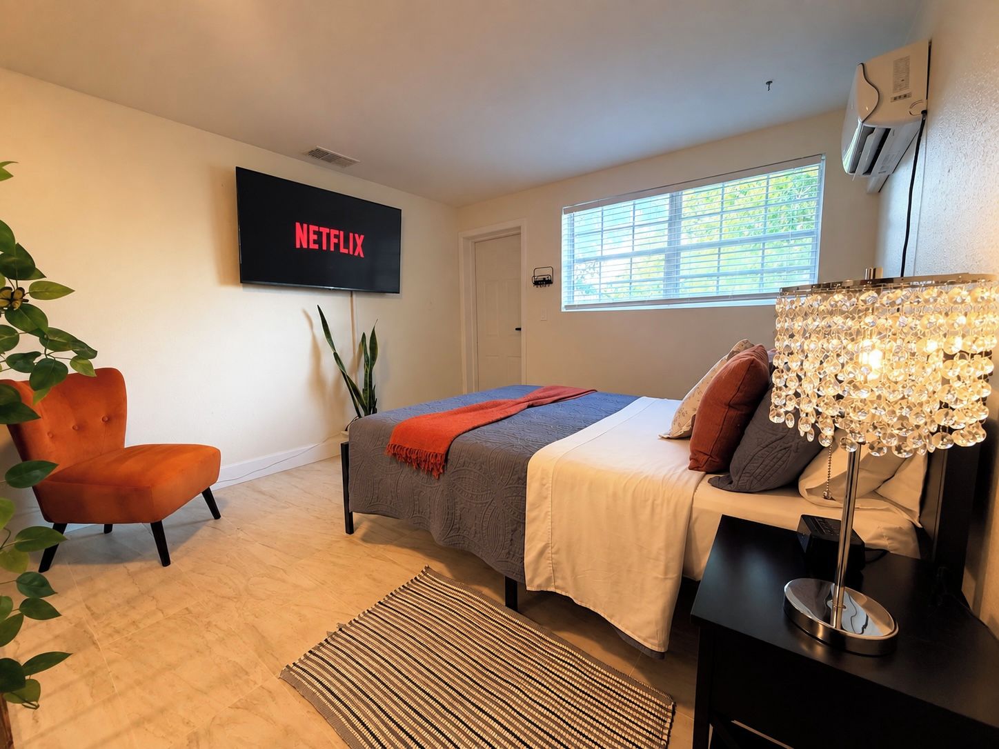 Ein modernes Schlafzimmer mit einem Bett, einem an der Wand montierten Fernseher, einem Fenster mit Jalousien und einer Pflanze.