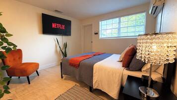 Ein modernes Schlafzimmer mit einem Bett, einem an der Wand montierten Fernseher, einem Fenster mit Jalousien und einer Pflanze.