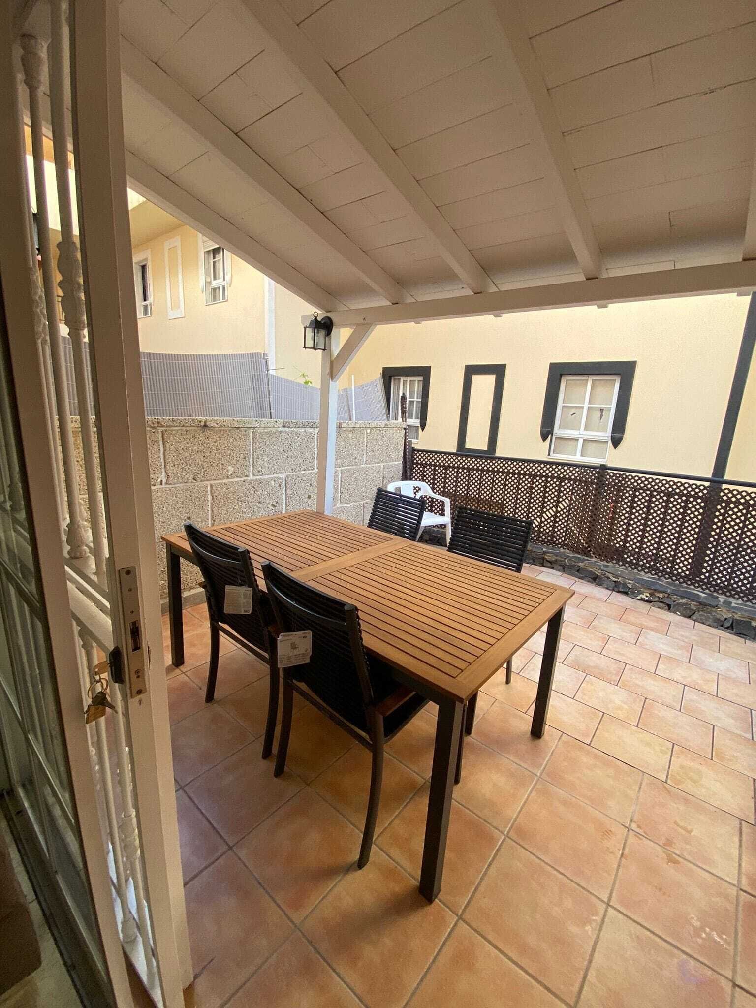 Une terrasse extérieure avec une table et des chaises en bois, un mur en pierre et un espace couvert doté d’un sol carrelé.
