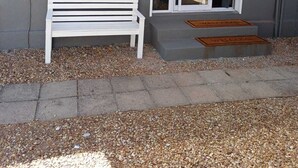 Un banco blanco frente a un edificio con pared gris, una puerta con paneles de vidrio y un felpudo.
