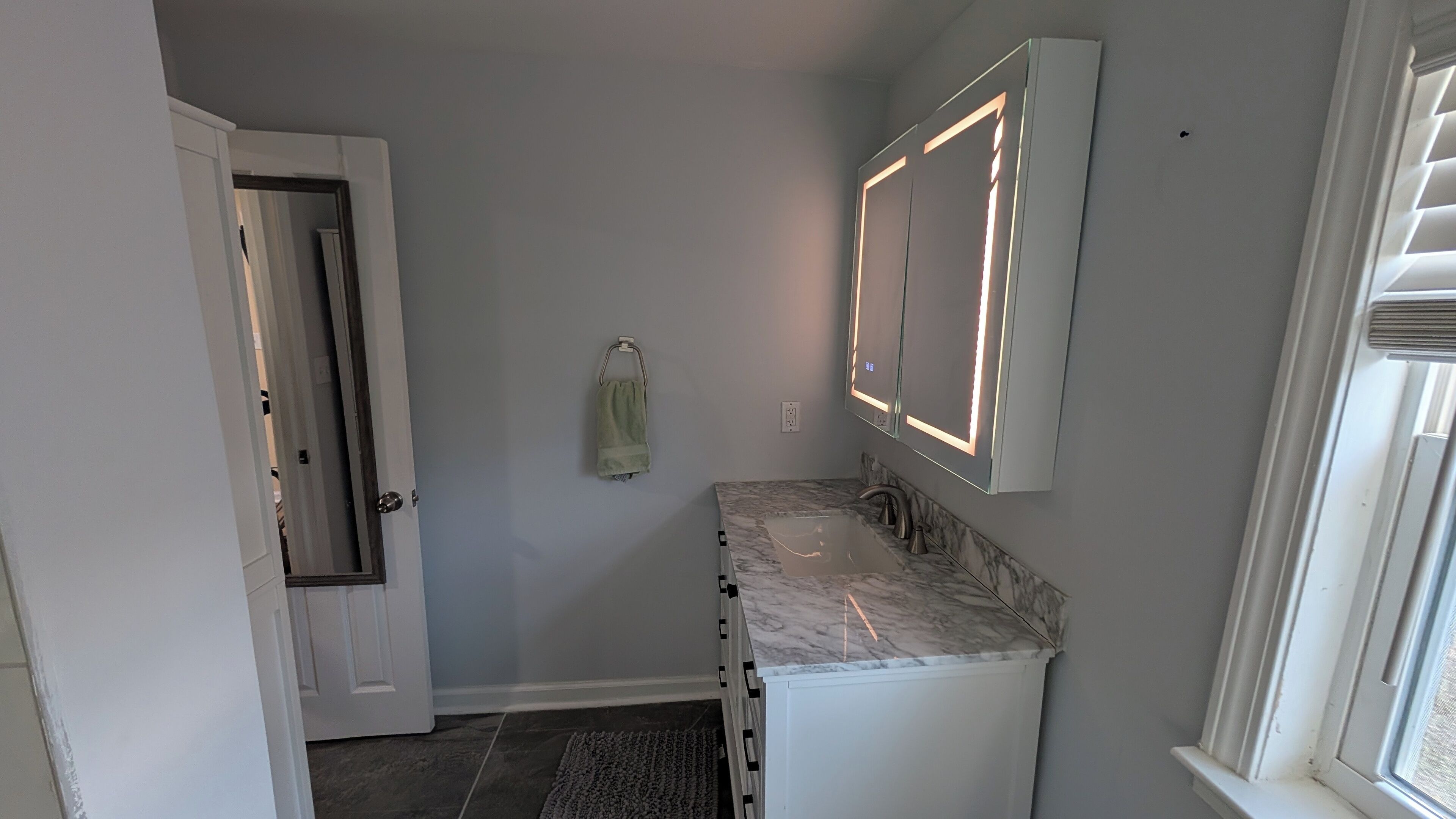 A bathroom with a marble countertop, a sink, a mirror with illuminated edges, a towel rack, and a door leading to another room.