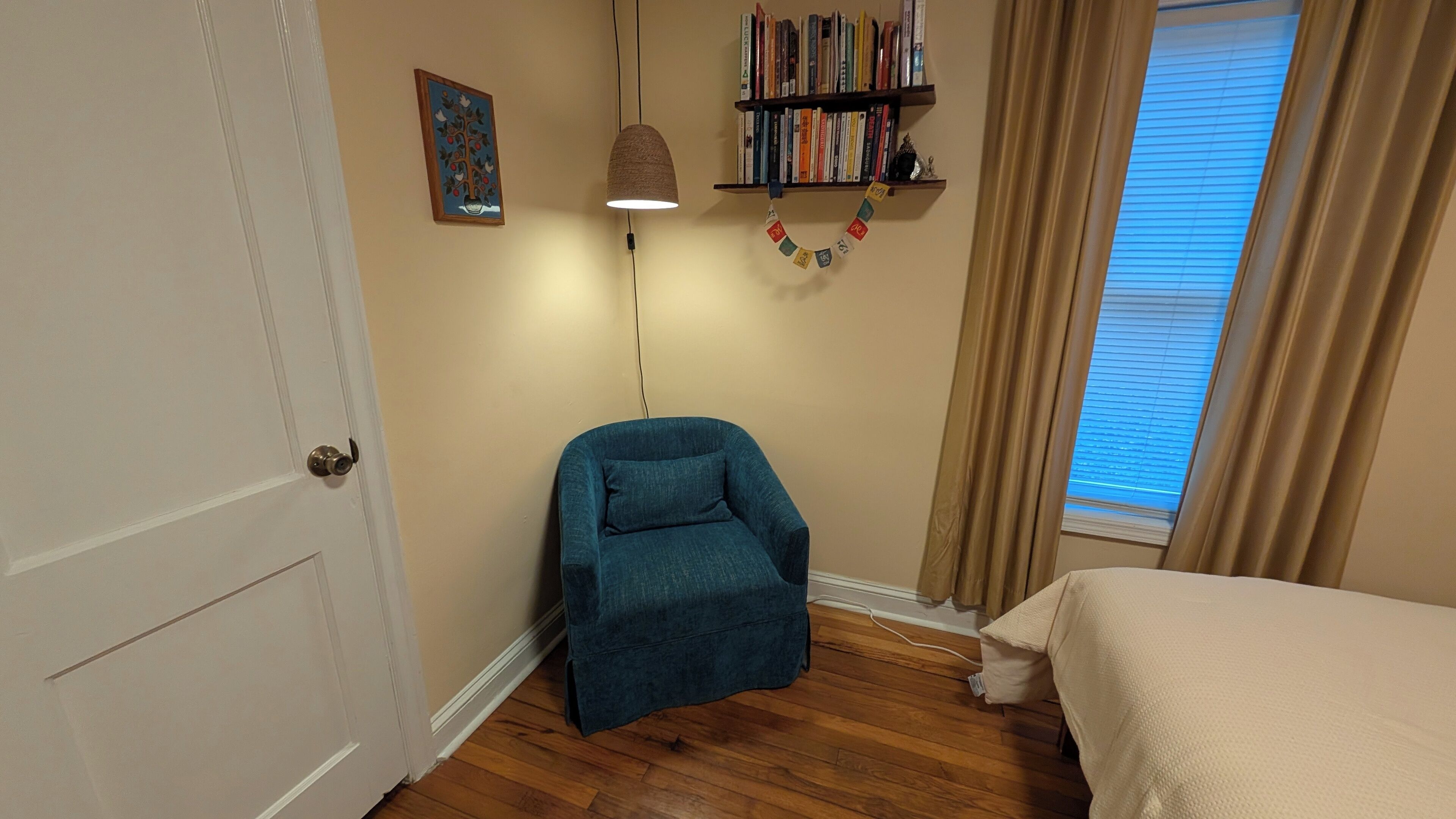 Una stanza con pavimento in legno, una poltrona blu, un letto, una libreria, una lampada a parete e una finestra con tende.
