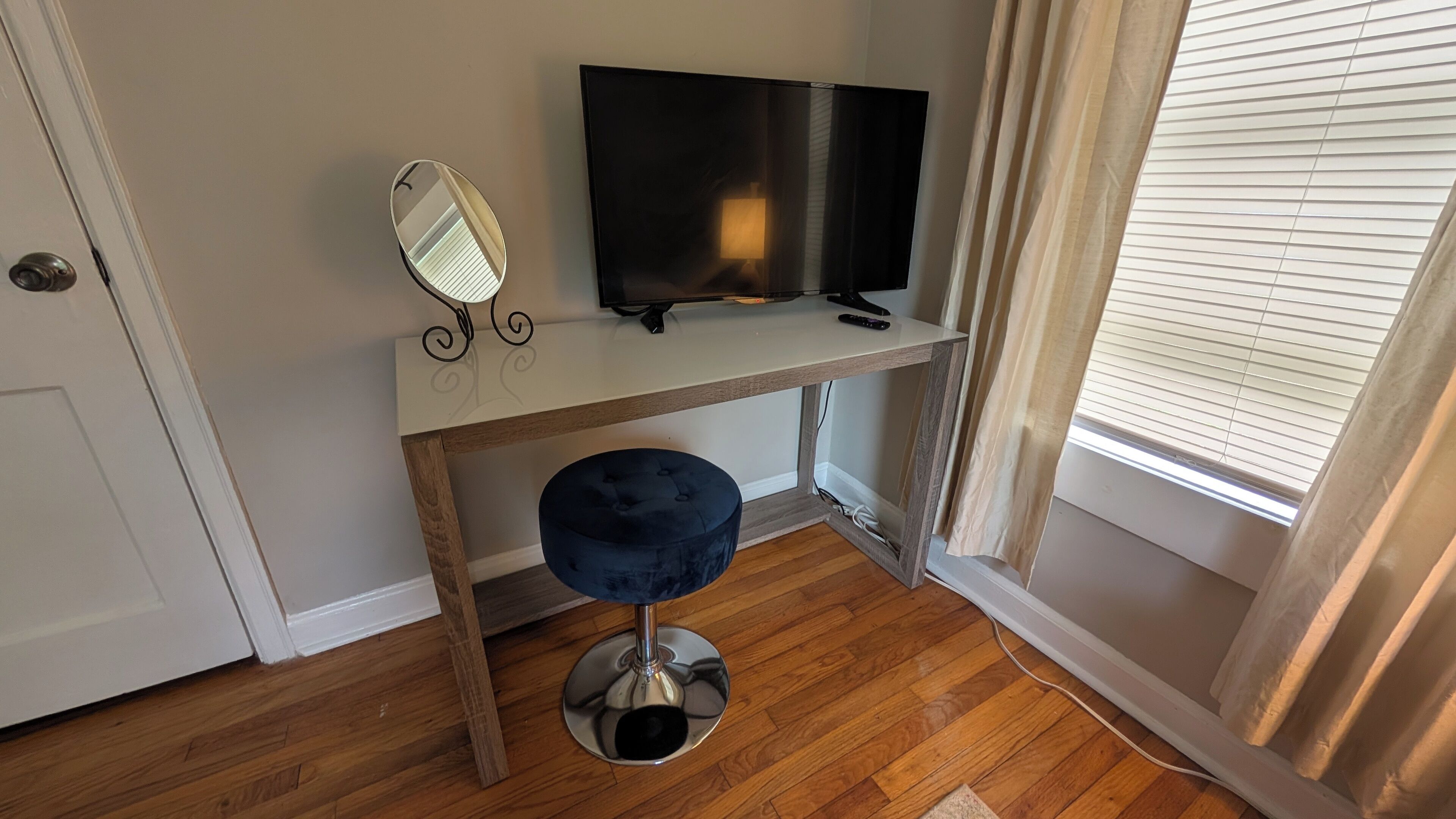 A room with a wooden floor, a white desk with a TV, a mirror, and a blue ottoman.