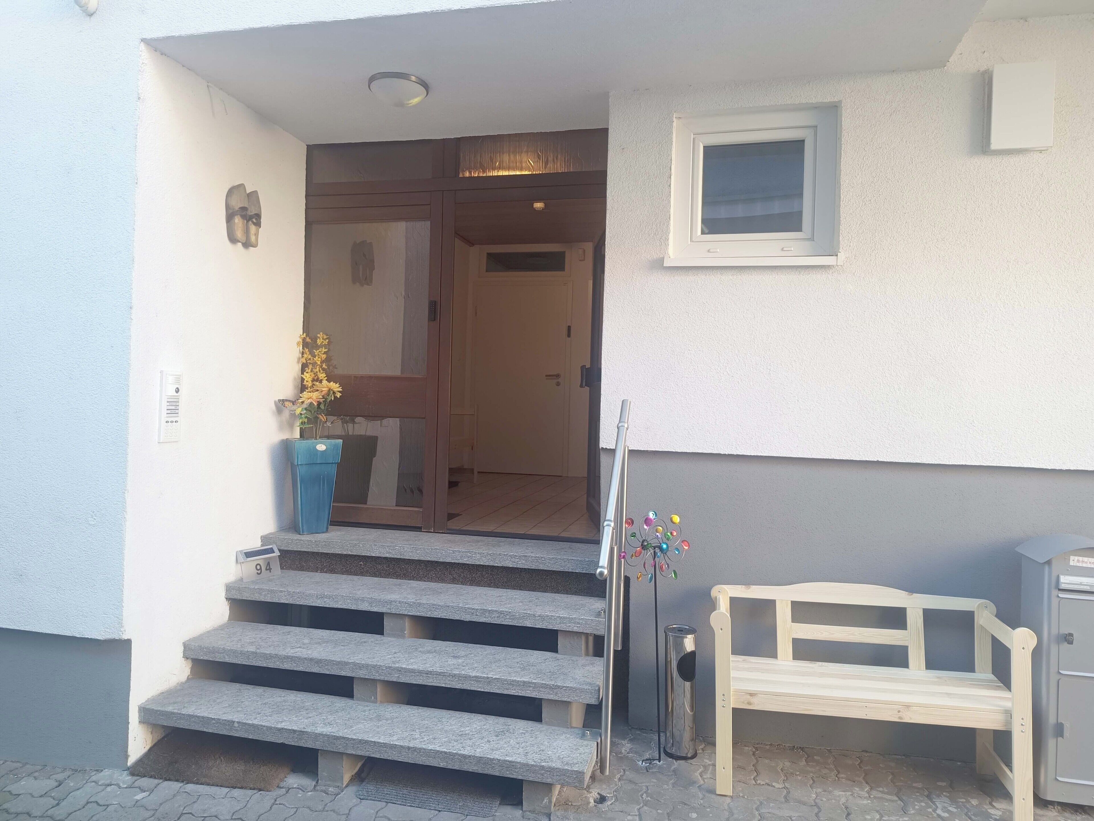 Une entrée d’immeuble avec un escalier en pierre menant à une porte, un bac à fleurs bleu contenant des fleurs jaunes, un banc en métal et une petite fenêtre.